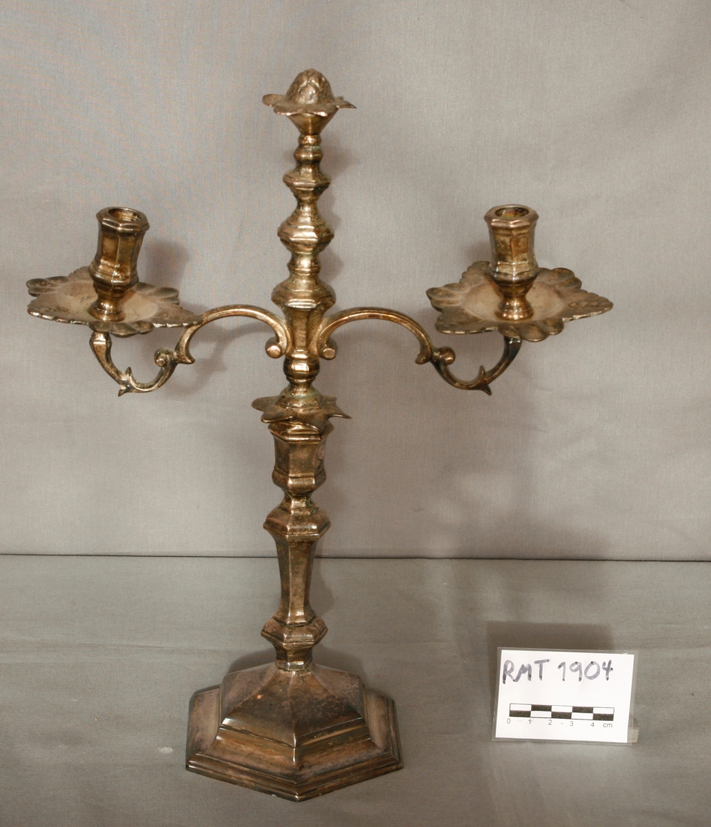 Toarmet stake med holdere til to lys. I midten en blomsterknopp. Kopi av lysestake i sølv. (Originalen er fra ca. 1710.)