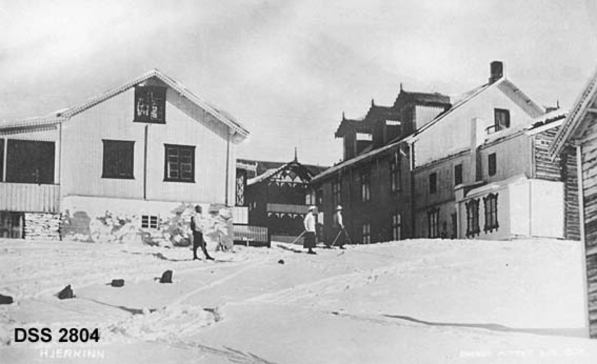 Våningshus på Hjerkinn fjellstue på Dovrefjell.  Sveitserstilbygninger med bolig- og utleierom.  Tre skiløpere står i tunet.  I forgrunnen til venstre stikker noen stabbesteiner opp av snøen. 