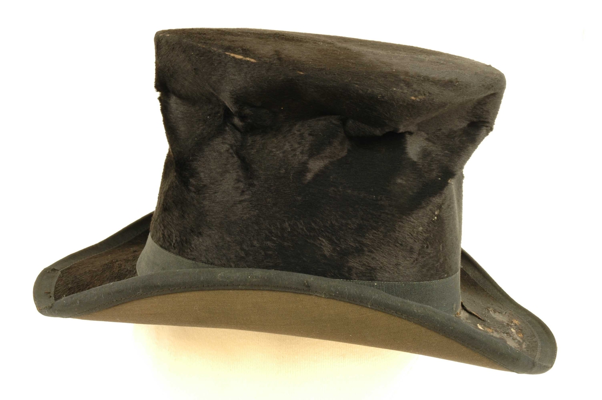 Svart flosshatt med høy pull og  oval bred brem. Svart silke hattebånd. Hatten er foret med silkefor, og bred innvendig skinnbesetning.