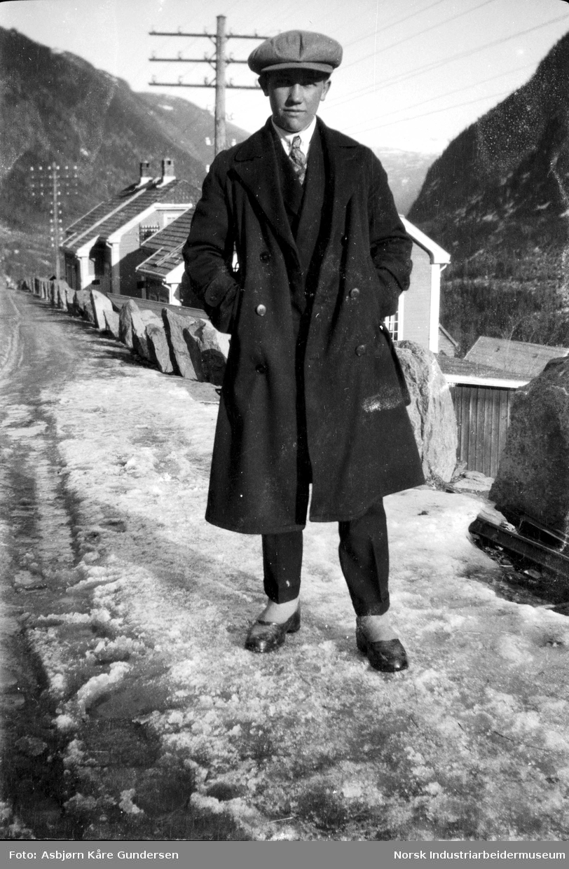 Ung mann med frakk, slips og lue i veien ved Våer. Bolighus og kraftledninger i bakgrunnen