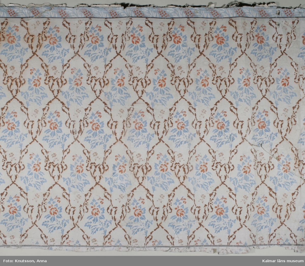 KLM 39750. Tapet, schablontapet. Schablonmålad tapet med vit bakgrund, röda blommor, blått bladmönster och brunt snedrutemönster. Ljusblå bård med röda blommor och vita blad bredvid varandra. Datering, 1840-tal.