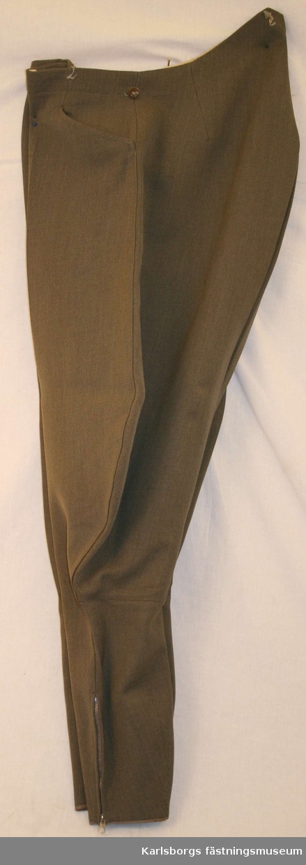 Ridbyxa m/1939 av gråbrungrön komiss. Två snedställda sidfickor en bakficka med en knapp. Dragkjedjor i byxbenens nederkanter.