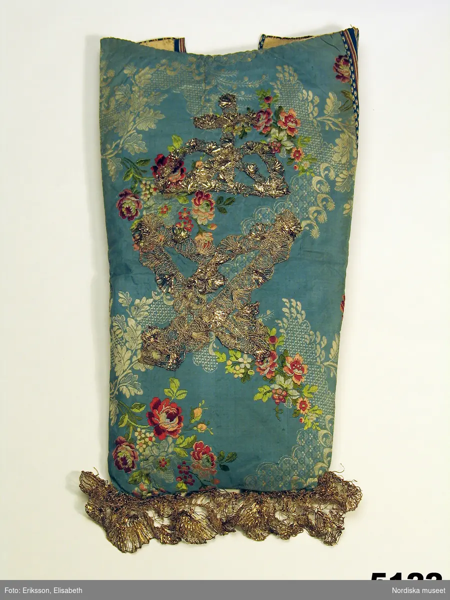 Doppåse av ljusblå sidenbrokad med rosenmönster i rött, rosa, grönt och vitt och invävda spetsband i ett mönster från 1760-70-talet.
Påsen liten och avsmalnande mot botten. Påsydd dekor av silverspets formad som en krona över ett hjärta? Nedtill kantad med en bredare guldspets. Ryggstycke av bomullslärft  med tryckt mönster med små fröliknande figurer ordnade i stramt randmönster i blått, brunt och vitt. Helfodrad med linnelärft, sprund i ryggen.
Kommer från Truls Bengtsson.
Enligt bil. "Minst 100 år gammal /1874/ men begagnad ända till 1830-tal".  Dock är inte bakstyckets bomullstyg så gammalt varför man kan tvivla på uppgiften.

Berit Eldvik febr 2006