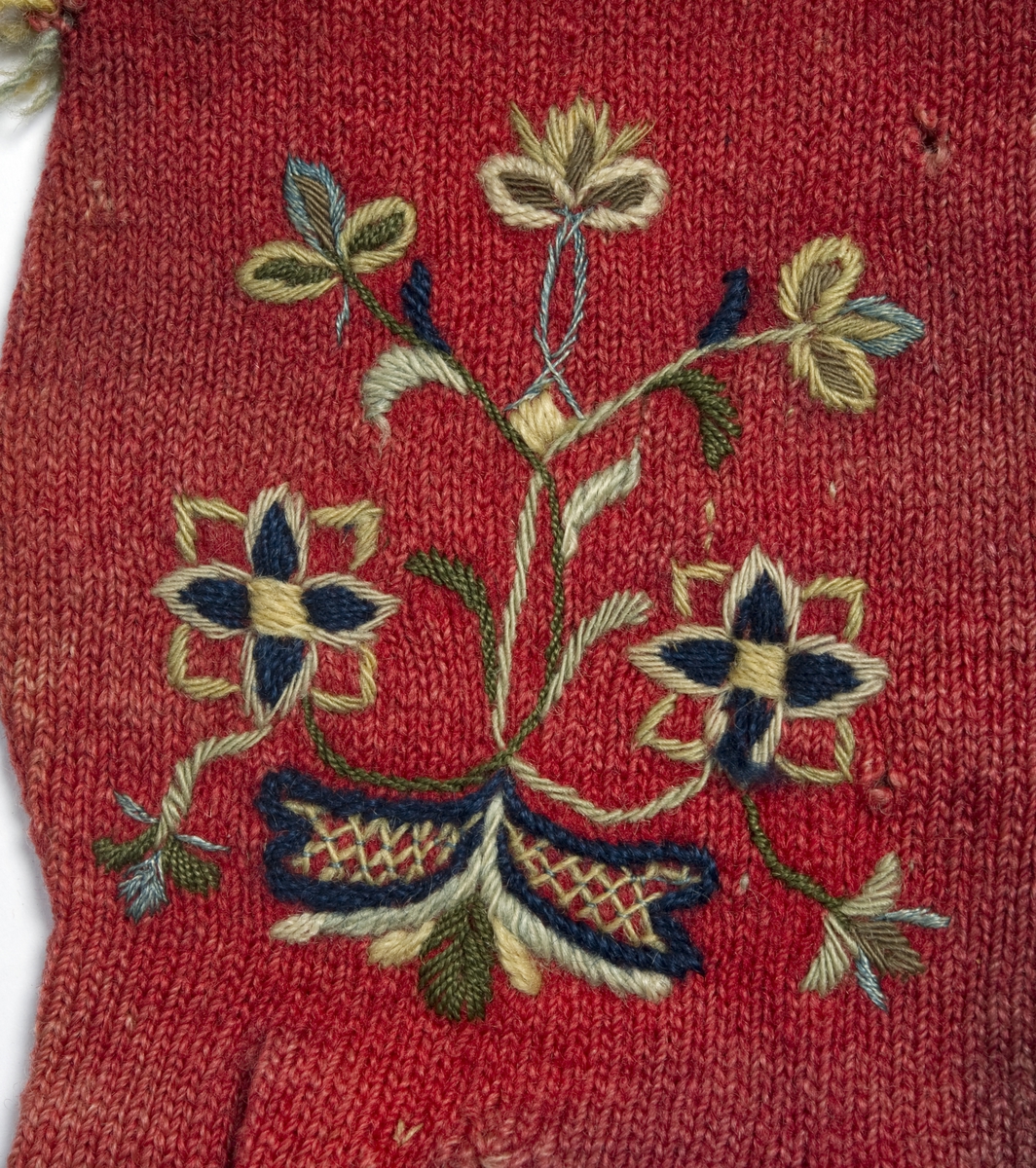 Rød strikket fingervante av ull, dekor av broderi og frynser.