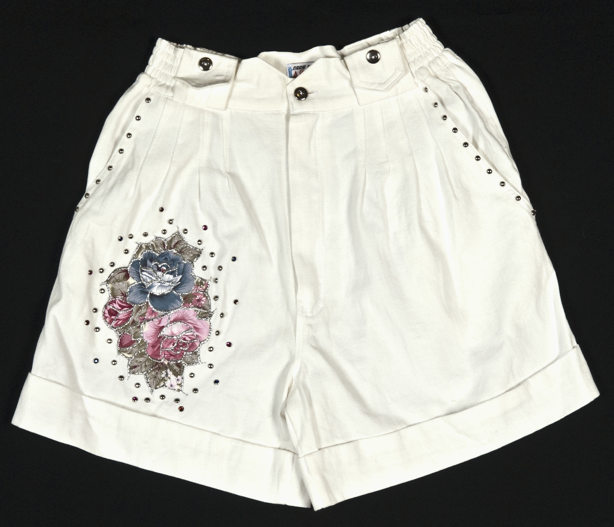 Hvit jakke i bomull med dekor av kuler og fargestener med applikerte felt av blomstermønstret tekstil med glitter på. Tilhørende shorts med samme farger og dekor. 