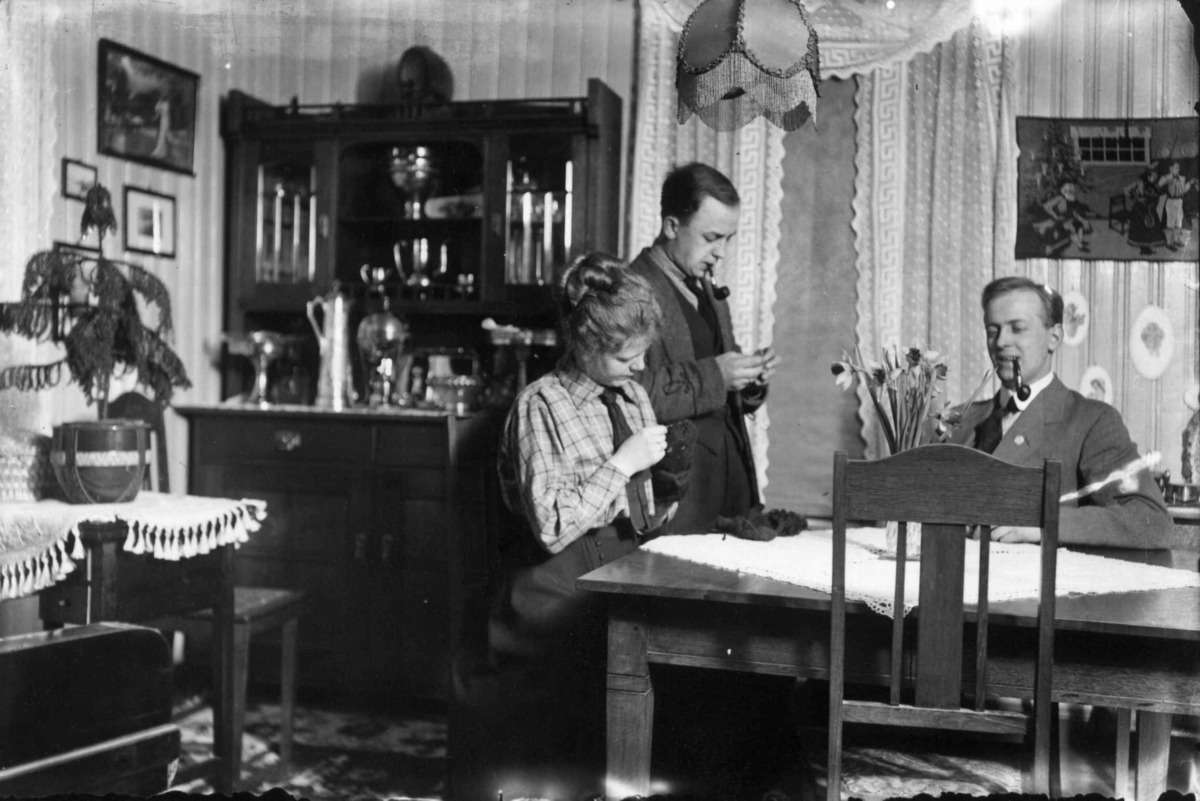 Stueinteriør. En kvinne og to menn ved spisebord, buffet, potteplanter, gardiner..Trolig Fredenborgveien 2, Oslo, ca. 1916.