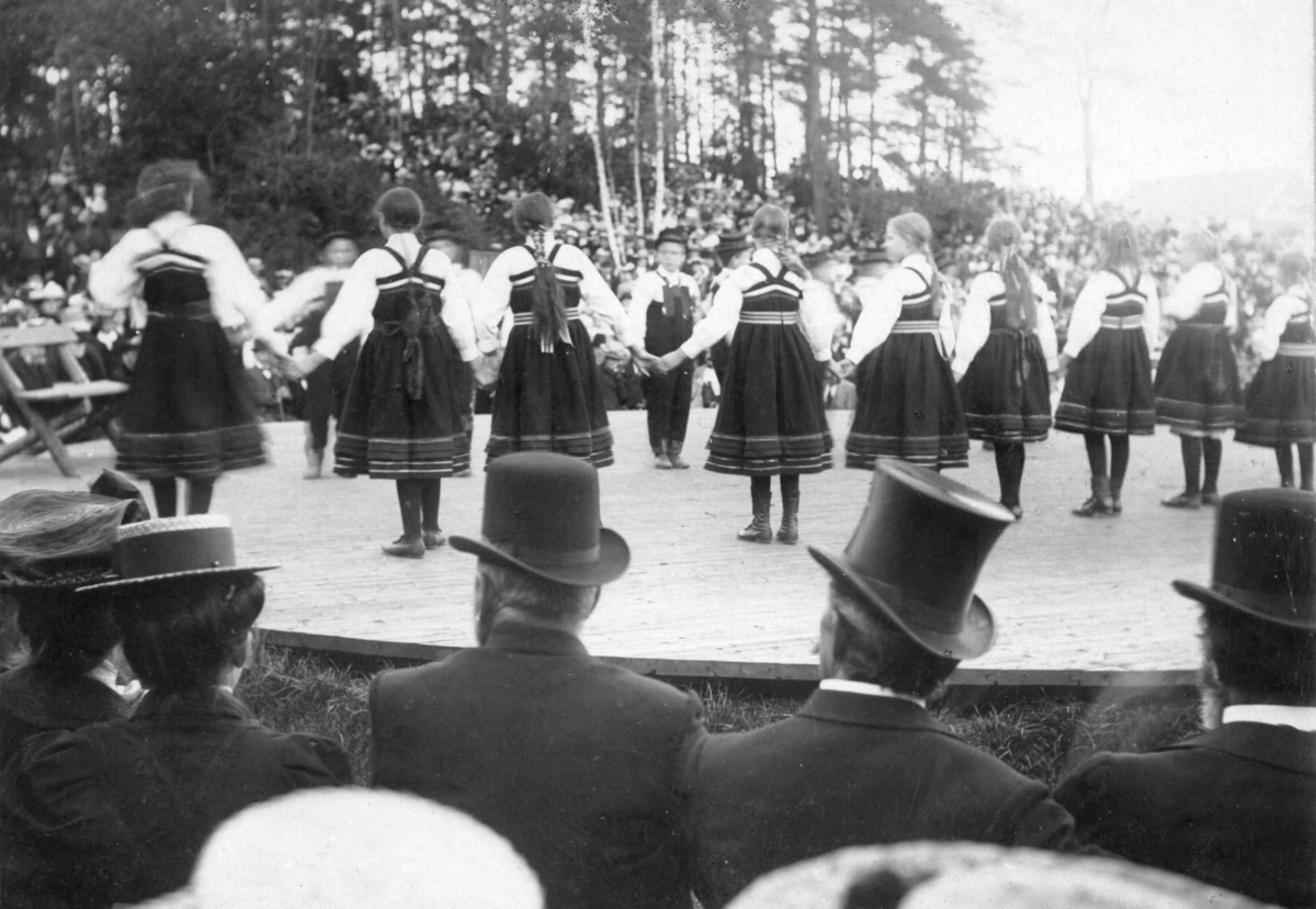 Høstfest på Norsk Folkemuseum i 1902. Folkedansgruppe på scenen. Publikum med hatter og flosshatter i forgrunnen.