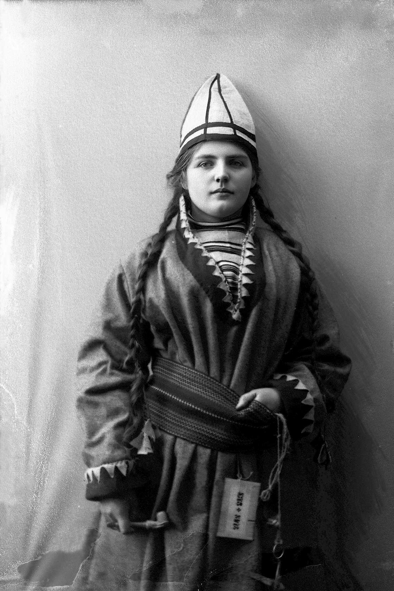 Kvinne i samisk inspirert drakt - kofte, lue og nålehus med beltering. I den ene hånden holder hun en pipe. Beltestakk fra en norsk bunad.