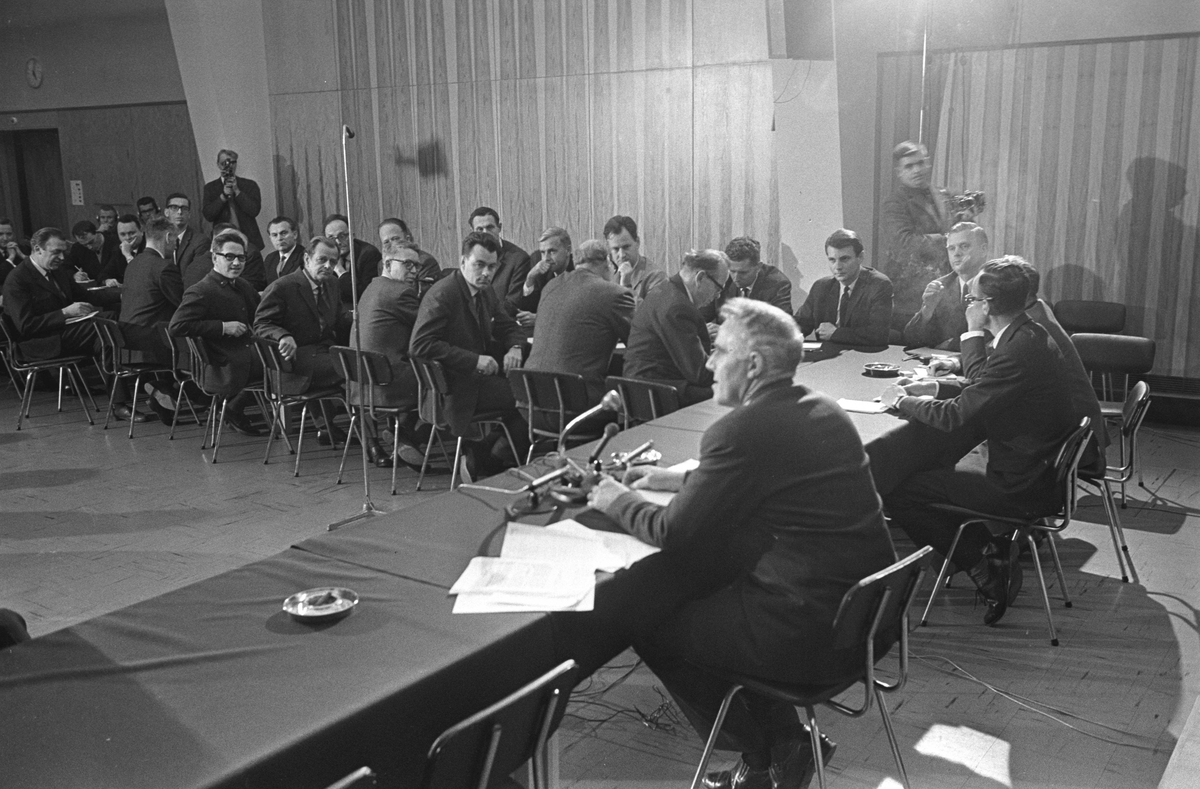 Statsminister Per Borten har pressekonferanse i den store møtesalen i den lave fløyen på vestsiden av Høyblokken i Regjeringskvartalet, oktober 1965. Bortens regjering tiltrådte 12. oktober 1965. Dette er derfor mest sannsynlig den første pressekonferansen etter tiltredelsen, holdt samme dag.