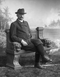 Portrett, mann i ytterklær på benk. Strømper og nikkers.