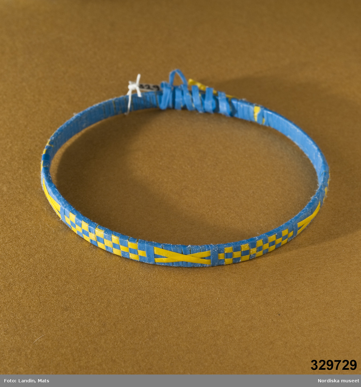 Runt armband med mönster vävt av tunna plastband. Mönster i form av gula/blå rutor och kryss. Afrikanska influenser.
Inv.nr 329.728-329.730 är likadana.
Anm.: Bandet har släppt på ett ställe.
/Zingoalla Rosenqvist 2009-02-05