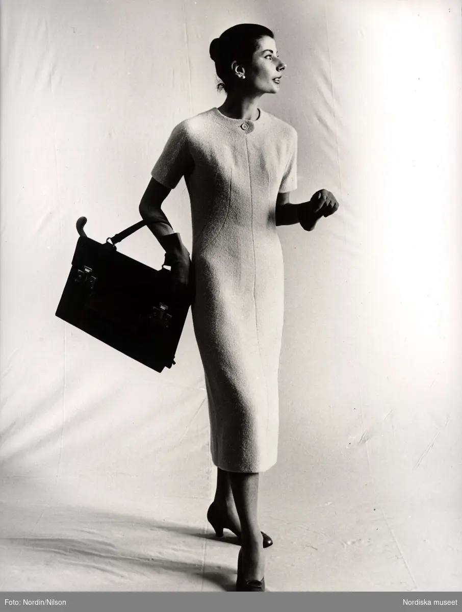 "Original Givenchy". Nordiska Kompaniet. Kvinna i figurnära klänning, handskar, pumps och med handväska.