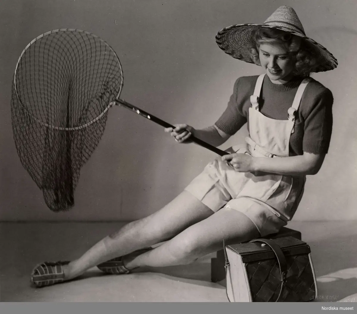 Sommarmode från varuhuset Nordiska Kompaniet i Stockholm 1944. En kvinnlig modell iklädd korta hängselbyxor, tröja, stråhatt och sandaler sitter med en fiskehåv i handen. Vid hennes sida står en väska av flätat näver.