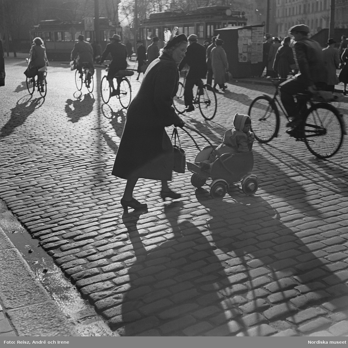 Kvinna med barnvagn. En kvinna skjuter en sittvagn med ett barn i framför sig, hon korsar just en gatstensbelagd gata där en rad cyklister passerar. I bakgrunden fler fotgängare, spårvagnar och en hållplats.