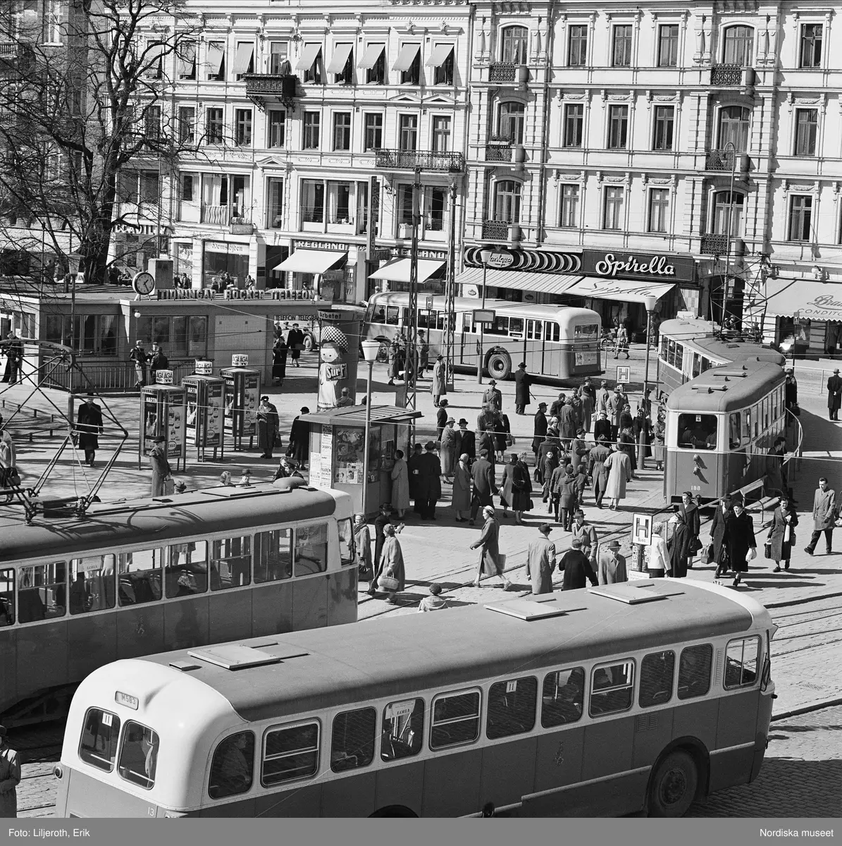 Vy över Gustav Adolfstorg i Malmö. En myllrande trafikbild med bussar, spårvagnar och fotgängare. I fonden husfasader med butiker, bland annat Spirella.