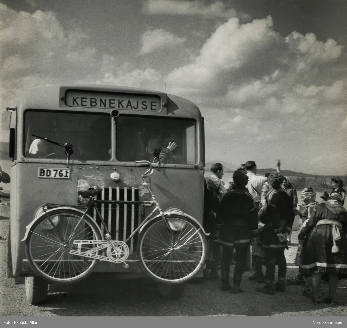 Samer och turister vid buss. Holmajärvi, Norrbotten. Bussen är skyltaad "Kebnekajse" och har en cykel monterad fram.