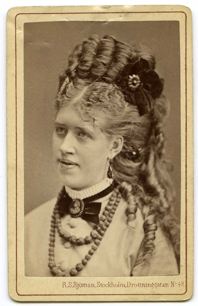 Porträtt av ung kvinna med hög, konstfärdig frisyr med en sammetsblomma som hårsmycke. Kvinnan har en vit skjorta med veckad ståkrage, sammetsband med en kamé runt halsen samt ett pärlhasband och hängande örhängen. 1870-tal.