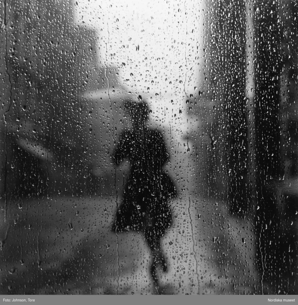 Gatuscen, Paris. Regndroppar på fönsterruta. I bakgrunden konturerna av en kvinna med paraply.