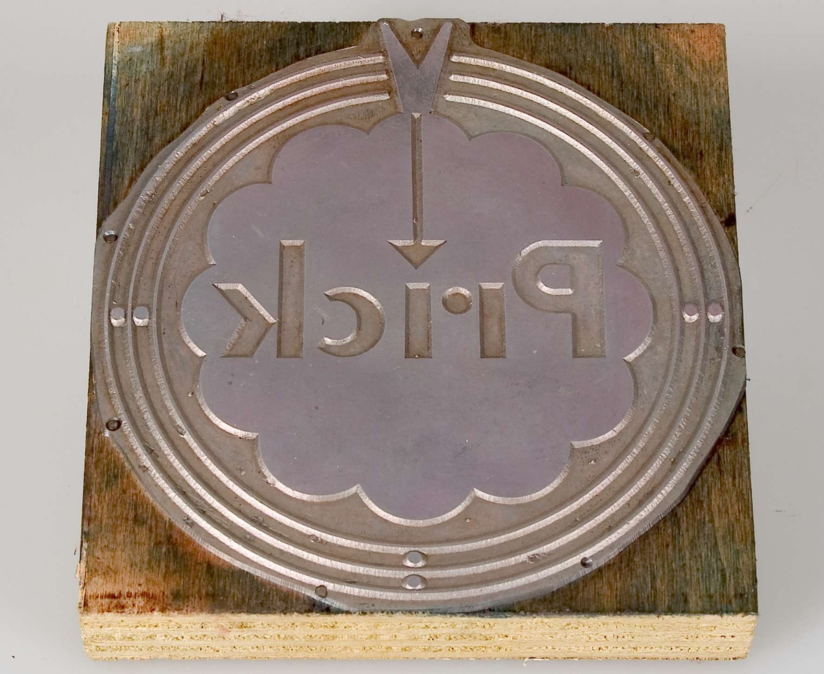 En kliché av metall, monterad på en kvadratisk platta av skiktlimmat trä. Rester av färg. Motiv: logotypen Prick.