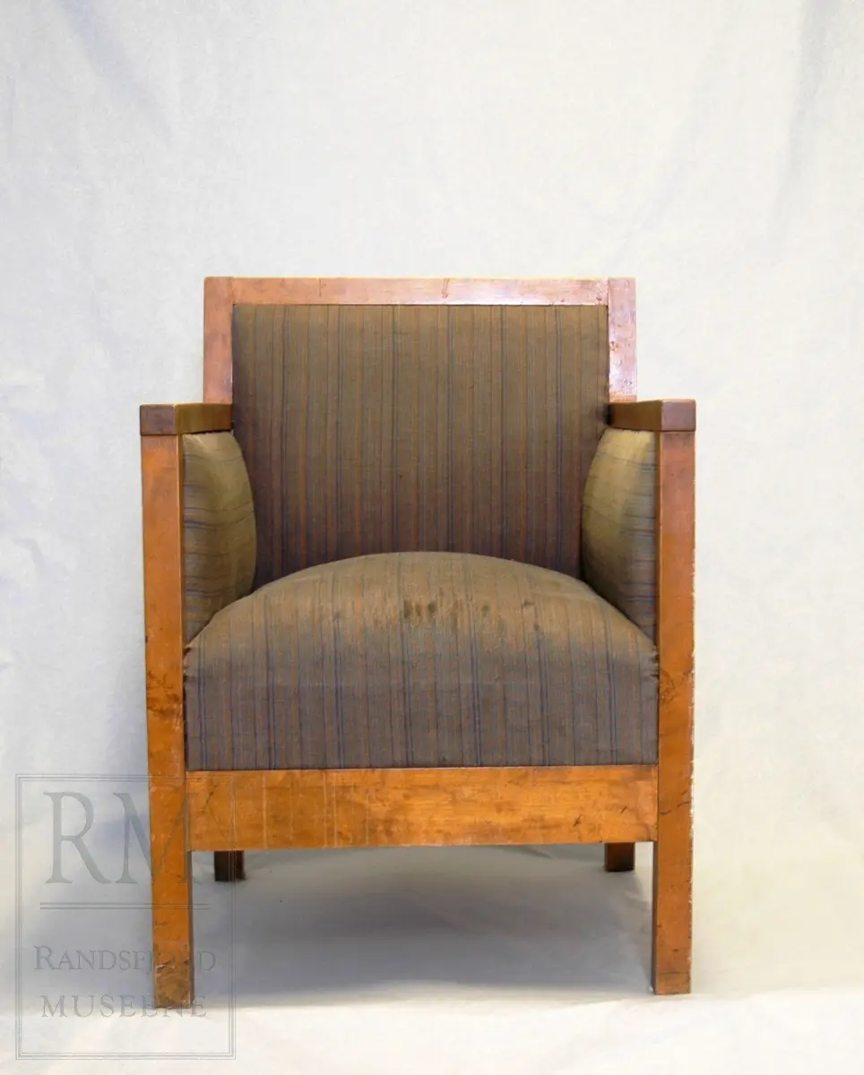 Lenestol med firkantede, korte bein.Stolen er som en kasse med rett rygg og høye armlenere.Yttersiden er i tre/finer, innsiden er polstret og trukket med stoff. Det er store fjærer i setet.