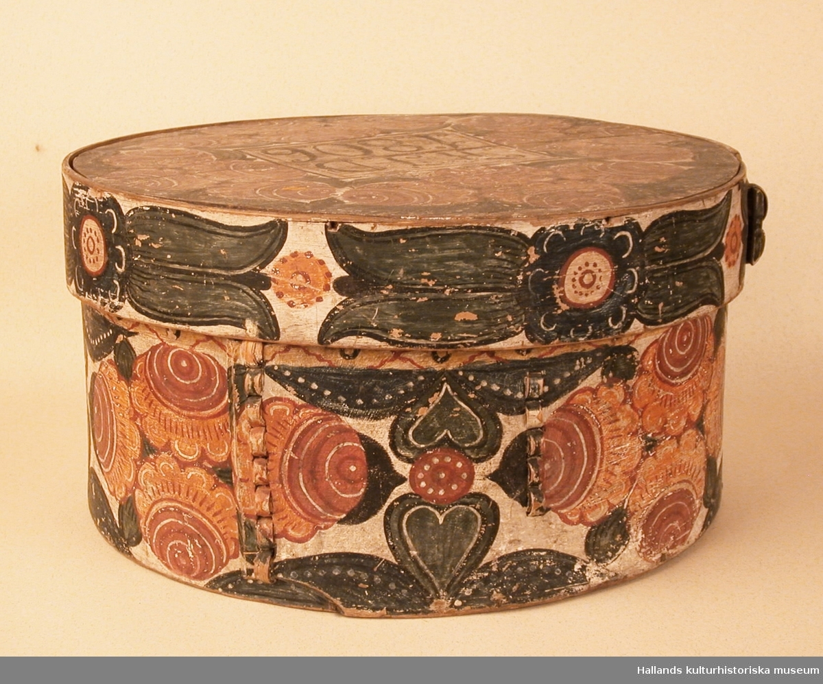 Cylindrisk, svept ask av björk, botten och lock av furu. Målat rosmotiv i rött och blått. Initialerna KJD samt årtalet 1806 på locket.