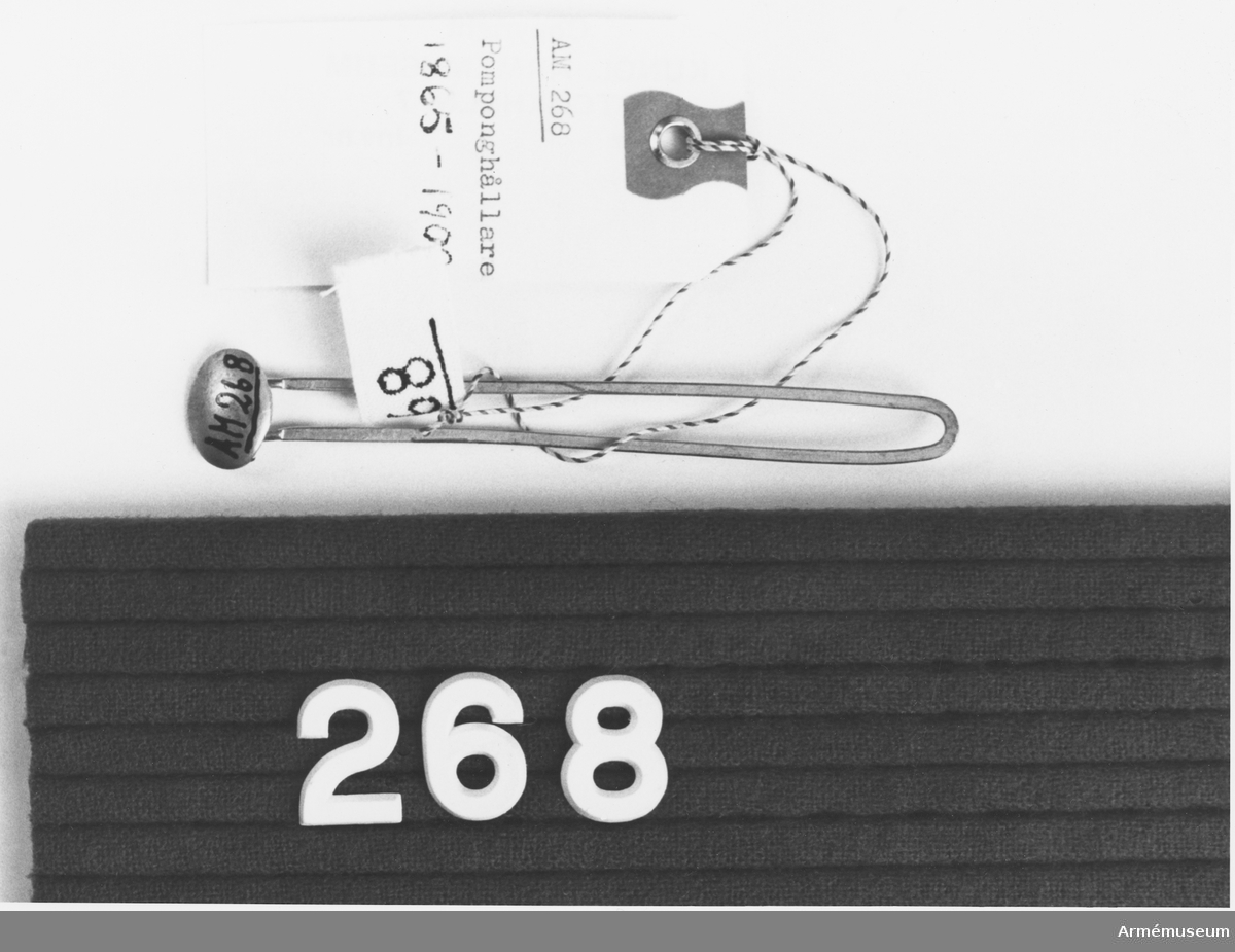 Pomponghållare m/1865-1908, I 5.
Rulla 1916. Består av en liten oval platta som skall tjäna som stöd åt pompongen. Dessutom en spärr för hållaren så att den sitter fast. Två skänklar utgår som ett utdraget "U" från  plattans baksida. Denna nål stickes genom pompongen, bakom vapenplåten och fasthålles med låsknapp.