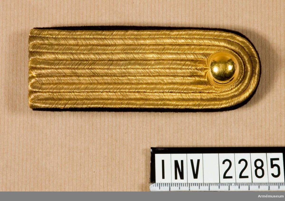 Fem rader guldgalon tätt bredvid varandra bildar överdelen av  klaffen. Knapp m/1901 för Karlskrona grenadjärreg. Foder och  hank av mörkblått kläde. Hake av gulmetall märkt "MEA,  Milit.Ekip.Akt Bolag".