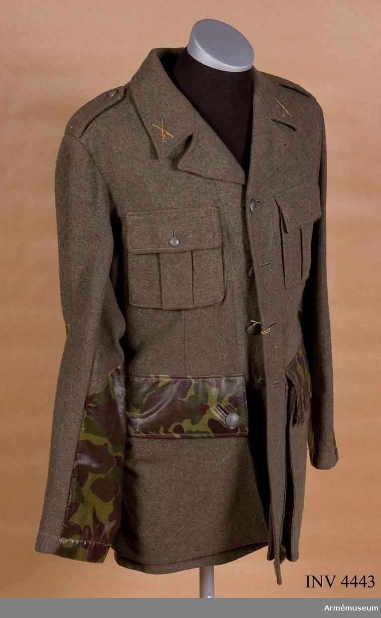 Vapenrock fm/1952 av kommisskläde med förstärkningar på ficklocken och nertill på ärmarna av kamouflagemönstrat vaxat tyg..
