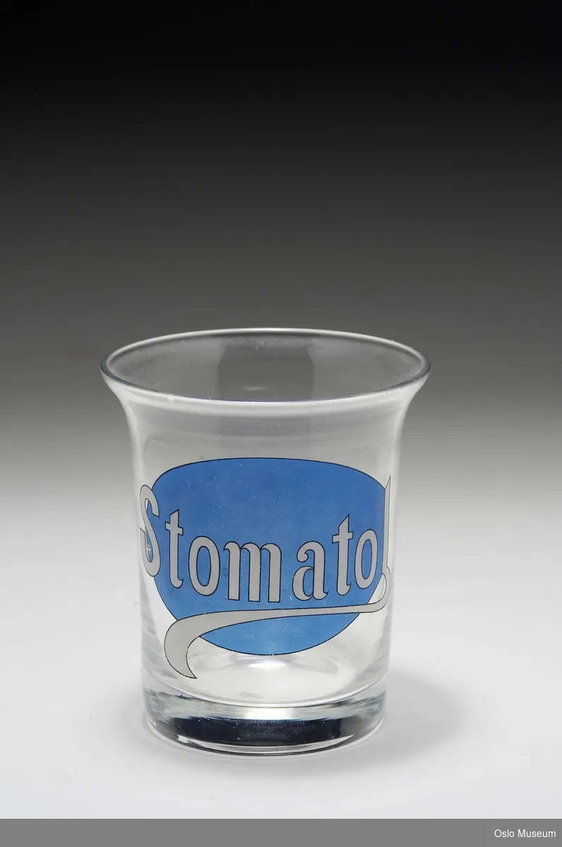 Sylinderformet klart glass, munningsranden lett utbrettet. Trykk på den ene siden av korpus; hvit tekst på lys blå bunn: Stomatol.