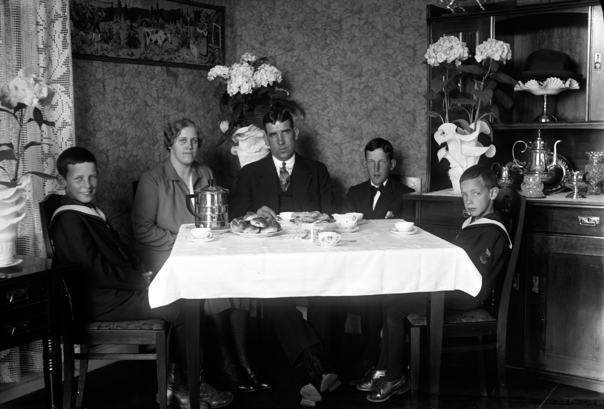 Kring kaffebordet sitter en finklädd familj. Det är dukat med vit duk, blommig servis och fat med bullar och kakor. Blommande Hortensior står i krukor klädda med kreppat papper. Observera hatten som ligger på ett fat i hyllan.