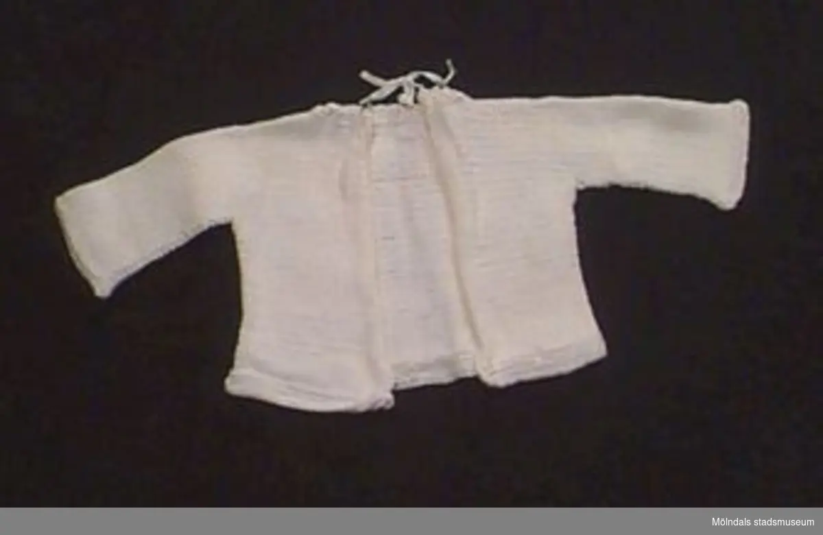 En vit stickad babykofta med öppet ryggparti. Knytning i nacken.