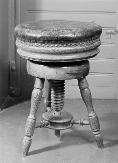 Stol, höj- och sänkbar med stoppad och skinnklädd dyna och tre ben. Stolen höjs och sänks med hjälp av en träskruv.