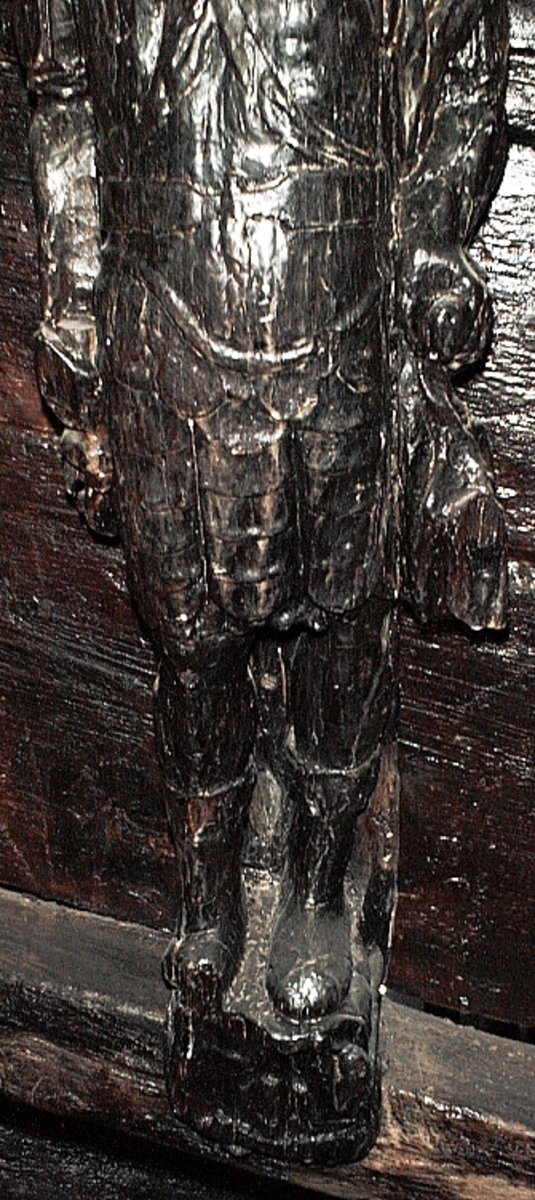 Skulptur föreställande en mansfigur med ett långsmalt och skäggigt ansikte, under en hög, slät hjälm med stora plymer. Runt hjälmen är en stålkrona trädd. Bakom huvudet skymtar ett snäckskal. Figuren är iförd en rikt ornerad rustning av thoraxmodell. Utefter figurens rygg hänger en mantel, som också draperar den vänstra axeln.

Hela figuren är frontalt framställd. Höger arm hålls lätt böjd utefter sidan, med handen fattande om ett svärdsfäste. Vänster arm är något böjd rakt ut från sidan, och handen verkar fatta tag om manteln. Kroppstyngden är jämnt fördelad på båda benen.

Figuren står på en sockel, vilken pryds av en bred och grimaserande maskaron. Sockeln är även försedd med en banderoll som tycks helt slät och utan namninskription.

Upptill är bakstycket framdraget i en kraftig "huvudplatta". Skulpturens baksida uppvisar tre snedställda, olika djupt liggande fält. Fälten, som utgör anliggningsytor, ger skulpturen en trappstegsformad profil.
Huvudplattan är delvis skadad. Svärdets klinga saknas. I övrigt är skulpturen relativt välbevarad.

Text in English: A sculpture representing a male figure with a long and narrow face that is bearded and has a moustache. On the head there is a plumed helmet on which a crown with points has been placed and behind the head a large sea shell. The figure is wearing richly decorated thorax type armour. The mantle is hanging down the back and draped over the left shoulder.

The figure faces towards the front. The right arm is slightly bent and held at the side and the hand holds a sword hilt. The left arm is slightly bent and held out from the side and the left hand seems to hold the mantle. The weight of the body is carried on both legs.

The figure stands on a plinth with a broad and grimacing mask and also has a label which is quite smooth and without any inscription.

The upper part of the back extends obliquely upwards in a powerful "head board". The sculpture''s back has three obliquely placed surfaces of different depths that are areas of attachment. The surfaces make the sculpture look like a step-ladder in profile.
The board behind the head is partly damaged and the blade of the sword is missing but otherwise, the sculpture is rather well preserved.