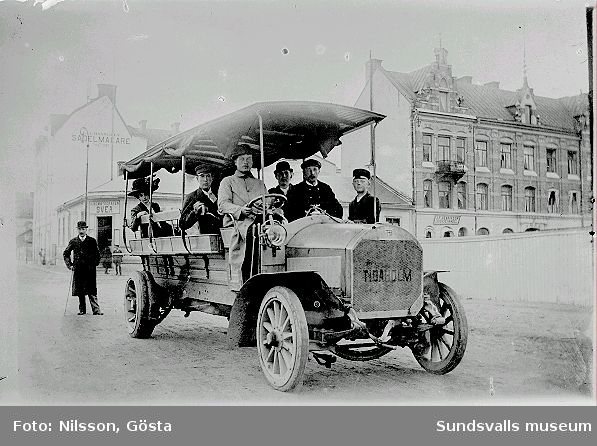 Den första omnibussen, Tidaholmsmodell. Föraren heter Axel Svensson och konduktören heter Erik Nilsson. I bakgrunden till vänster ses stadens näst första biograf "Svea" (f. d. Godtemplarloge).