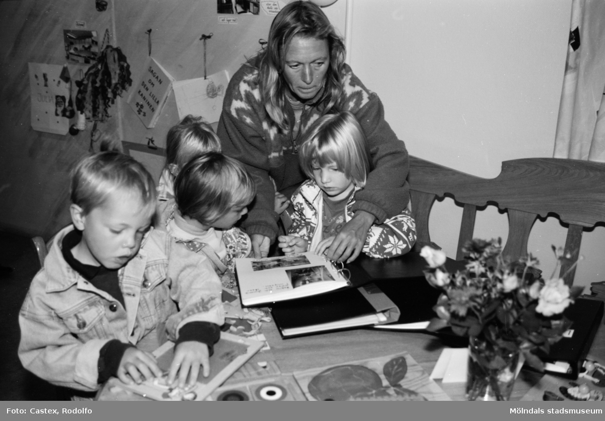 Fyra barn pysslar eller tittar i fotoalbum tillsammans med en vuxen person. Utställningsvernissage av och om Katrinebergs daghem på Mölndals museum 1993-09-10.