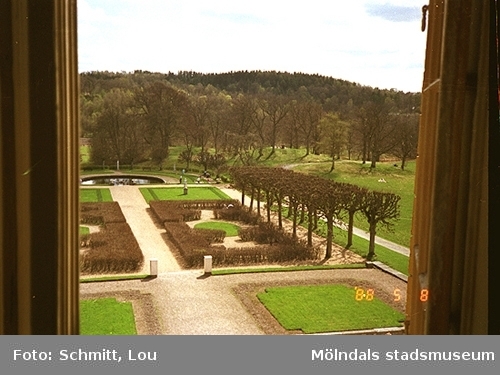 En del av parken på Gunnebo slotts högra baksida. Bilden är tagen inifrån och ut genom ett öppet brunt fönster. I bakgrunden skymtar man dammen.