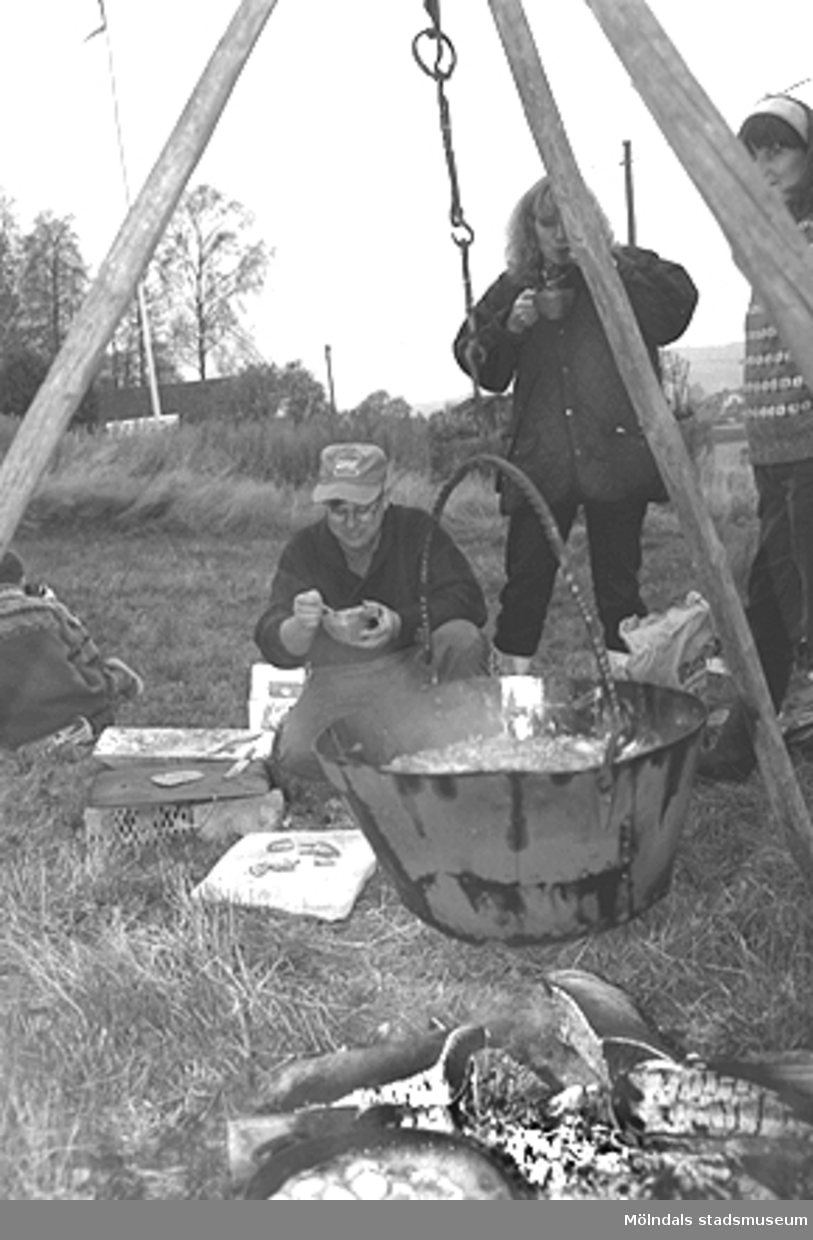 Familjesöndag vid Börjesgården, 1994-05-15. Närbild av en gryta som hänger över öppen eld. Sittande till vänster är museitekniker Alf Garthman och till höger står museichef Mari-Louise Olsson. De smakar på den färdiga grytan.