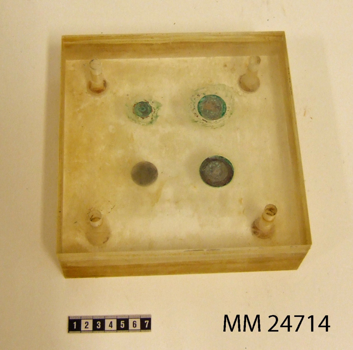 Platsblock av genomskinlig plast med fyra mynt ingjutna av valörerna 5 öre, 50 öre, 1 krona och 5 kronor. Hål borrat rakt genom plastkuben i alla hörnen med försänkning på ena sidan. Kuben delad och kan öppnas som med ett gångjärn.