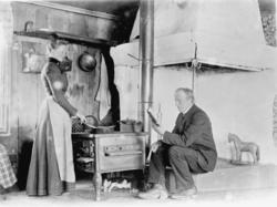 Kvinne lager mat på komfyren, mann sitter og leser i ei bok.