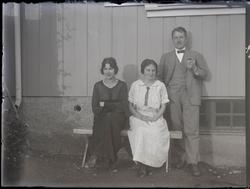 To kvinner sittende og en mann stående forran en trebygning.