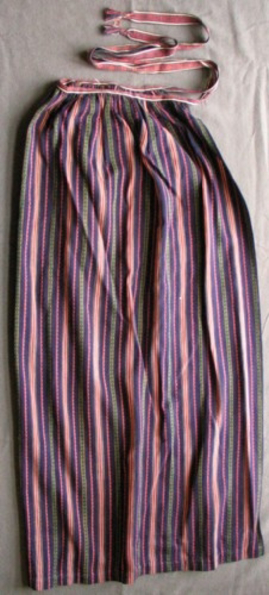 Rosengångsförkläde vävt i bomull med mörkblå botten, smala ränder i rosa/vitt samt rosengångspartier rosa/mörkblått och grönt/mörkblått/gult. Bredd nertill 895 mm, rynkat upptill vid linningen som består av ett band i samma mörkblå, rosa och vita bomullsgarn som i förklädet, dubbelvikt över linningen (10 mm syns), sedan slätt som knytband 660 mm (höger) resp 745 mm vänster. Avslutning med två flätor och tofs inräknat (40 mm). Fåll nedtill 20 mm, sidorna 4 mm. 
 
Eventuellt från Helsingborg enligt notering.