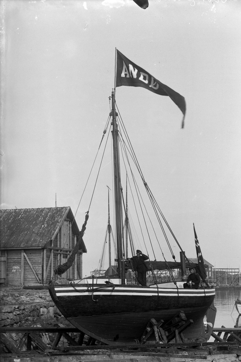 Lite seilfartøy på slipp. To mann poserer ombord, og båten fører både norsk flagg og en vimpel det står "Andø[...]" på. Bildet er antagelig tatt på Andenes.