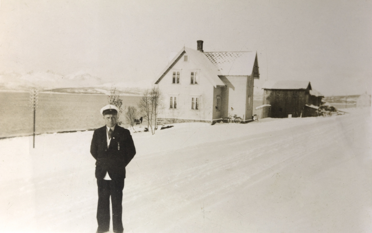 Ung mann med sangerlue, studentlue eller sjåførlue fotografert på en vintervei foran et hus ved en fjord eller sund. Kan det være i  Kvæfjord eller i Tjeldsundet?