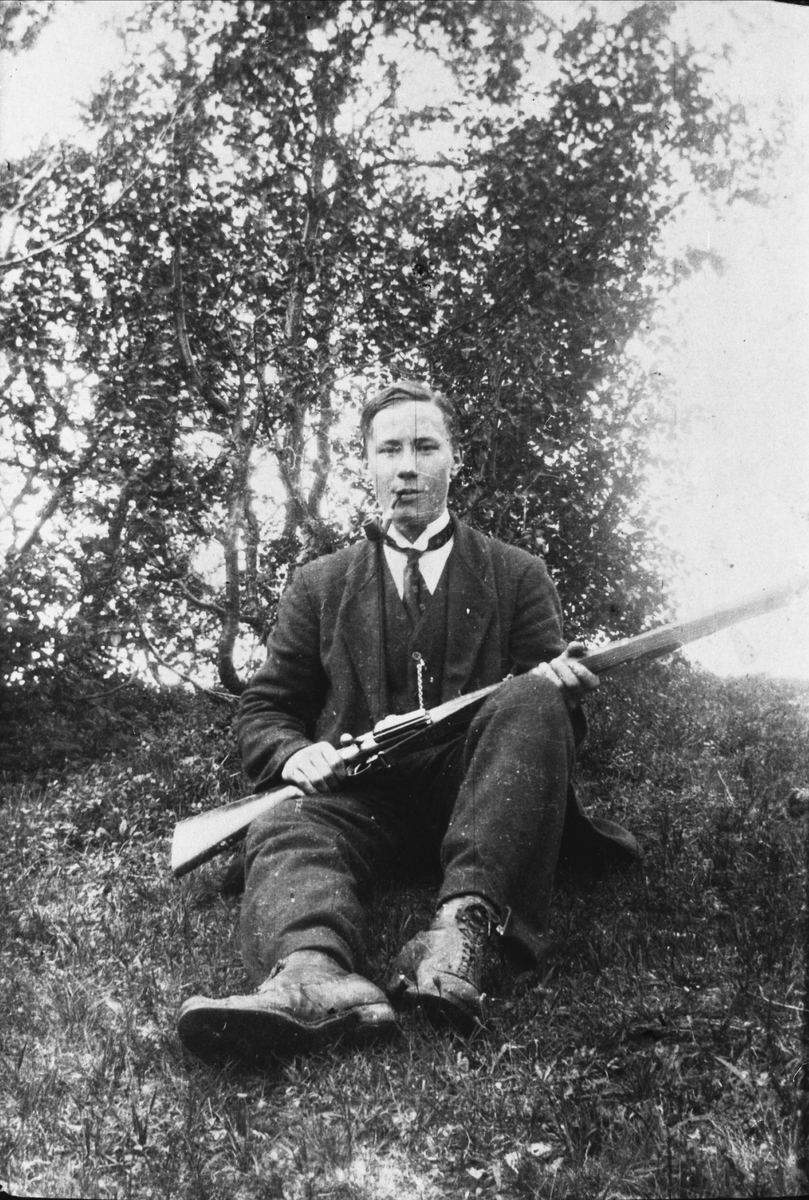 Ung mann med dress og pipe sitter ute på gresset med en Krag-Jørgensenrifle, antagelig rundt 1920 engang.