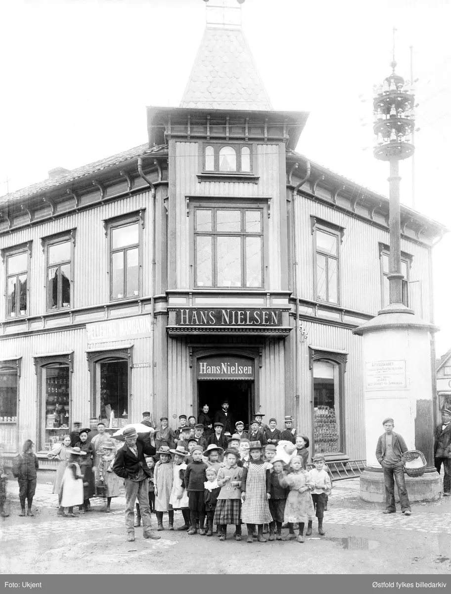 Hans Nielsens kolonialforretning i krysset Glemmengata og Brochsgate i Fredrikstad ca 1900-10. Adresse Glemmengata 1.
Gutten til venstre med trau på skuldra er ant visergutt i butikken, kanskje også gutten til høyre med kurv på armen. 
I døra ser vi kanskje butikkeier og kona, Hans Nielsen f.1860 og Marie f.1959, beggefra Skjeberg. Kolonialforretningen ble etablert 1886. 
Reklameskilt for Pellerins Margarin.
Stolpe for elektrisitet til høyre med skilt: 
"Aktieselskabet Fredriksstad Gas & Elecktricitetsværk
Leverer.... drivkraft for Fabrikanlæg."
Hushjørnet er del av det stedet man i Fredrikstad kaller "Det Glade Hjørne".