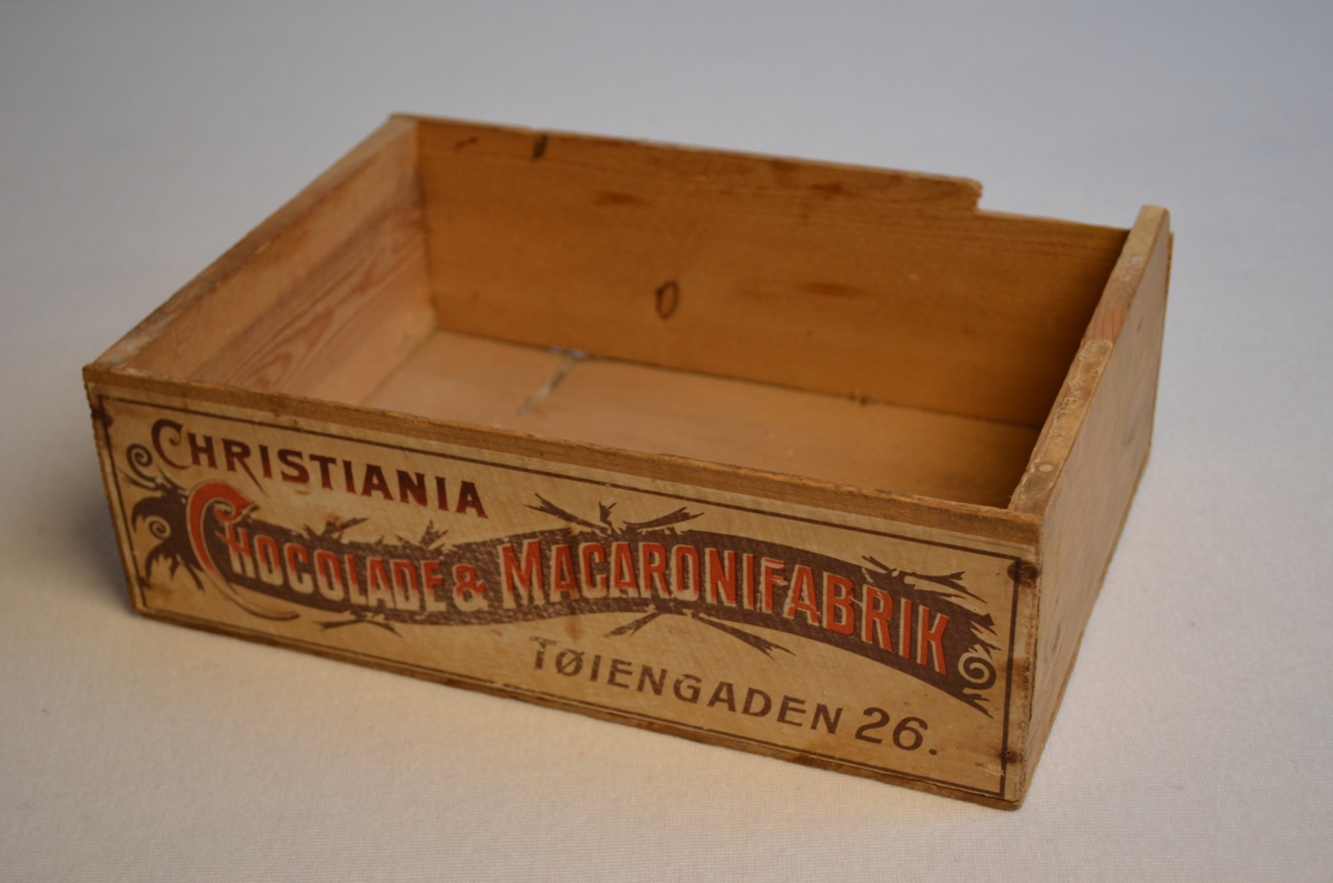 Lita trekasse utan lok som opprinnelig kom frå ein sjokolade og makaronifabrikk i Oslo. Ein del av kanten på kassa er broten av. Logoen på kassa er i raudt og brunt.