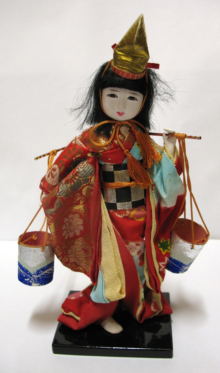Docka i traditionell dräkt som tillhör Vera Hanssons docksamling.

Köpt 1959. Yoriko. Japan.
Japanska i mångfärgade kläder huvusakligen rött.
Hon bär ett ok med två ämbar.