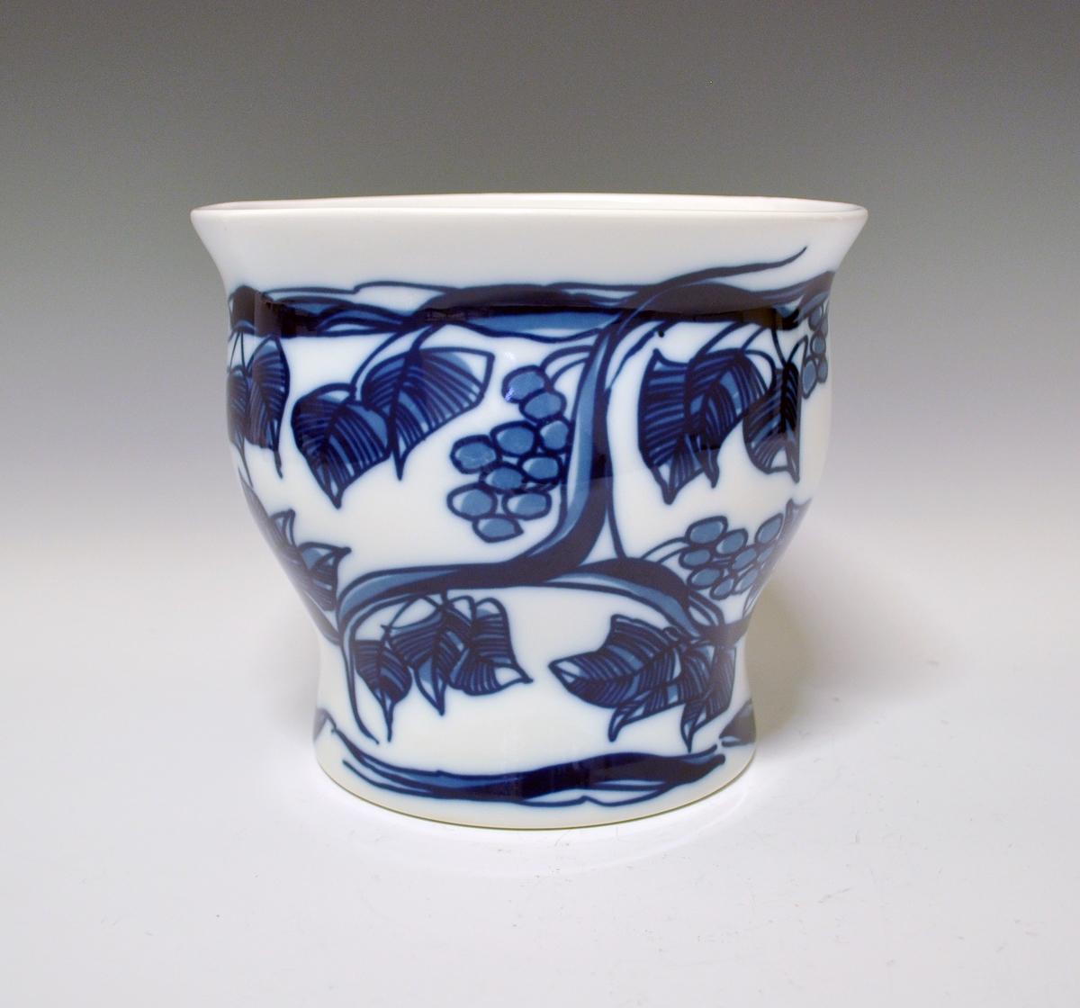 Vase i porselen. Bølget form, sirkelformet fot og oval åpning på toppen. Hvit glasur. Blå underglasurdekor: Grener med blader og drueklaser.
Kunstner: Unni M. Johnsen.