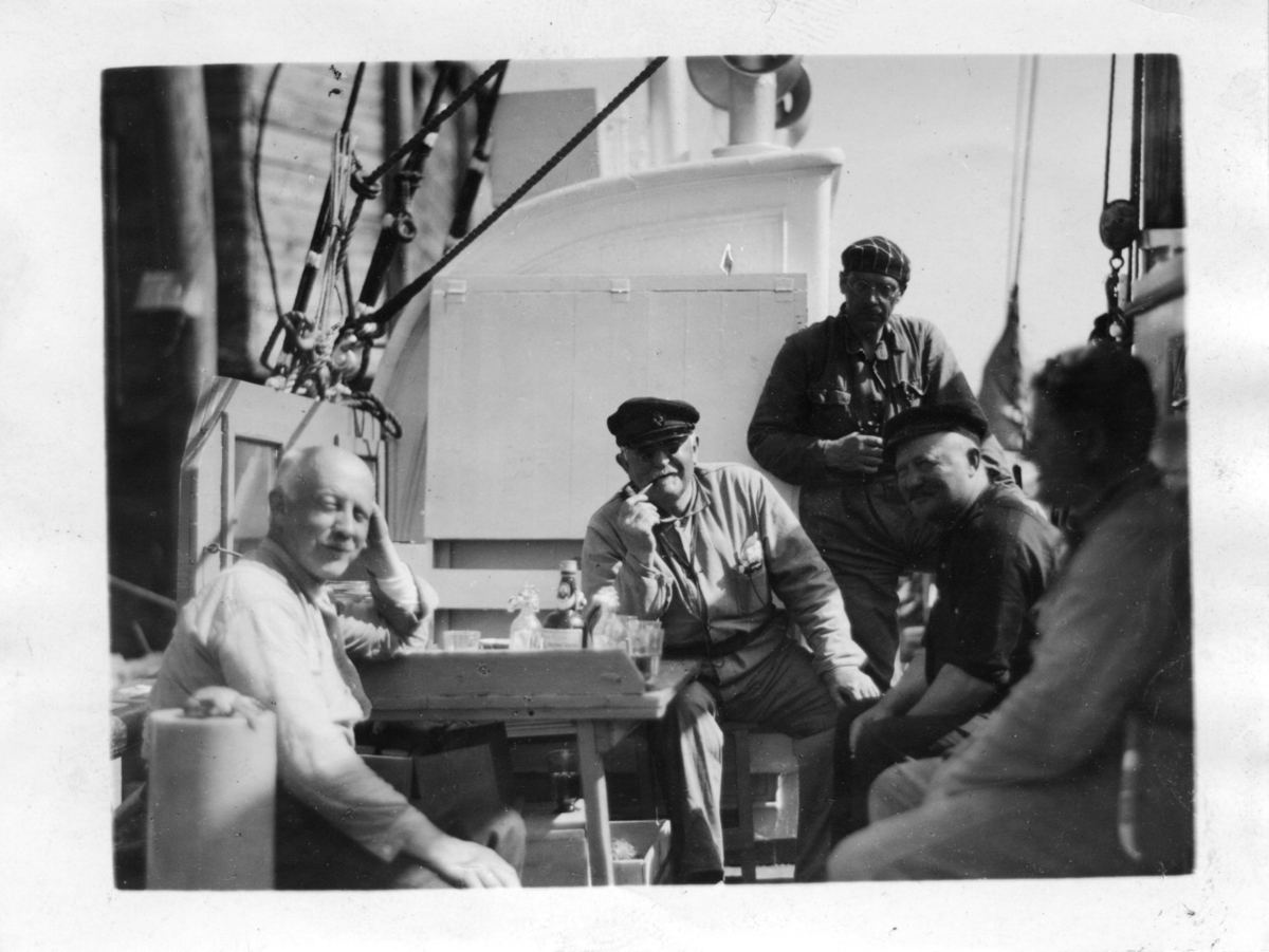 '5 män ombord på båten ''Gunnerus''. Andre från vänster Leonard Axel Jägerskiöld och stående till höger om honom är Nils Odhner. Andre från höger sitter C. A. Aanderud. Sittande vid bord dukat med glas och glasflaskor. På bilden även man vid namn Carlson. ::  :: Enligt  text på baksidan av fotografiet: ''bvalskasjouren (?) keskgeer(?) C. A. Aanderud (andre från höger) på visit ombord 'Gunnerus', Carlsons, Jägerskiöld, Nils Odhner(?) ska senare bestyra såväl i Norr som sydkustsvalfångst. J., som en sommar på 90-talet varit med, och Aanderud fann varandra helt.'' ::  :: Senare tillägg: ''Sune Swärd längst t. höger.'' ::  :: Ingår i serie med fotonr. 3679-3693 se även 3694-3719.'