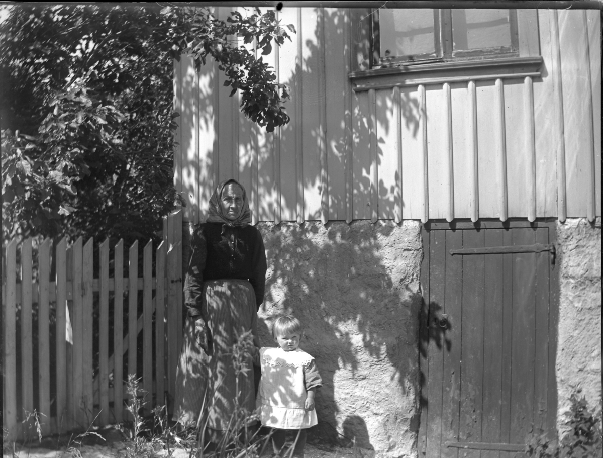 'Bildtext: ''Kyrkesund 1927.'' :: Vy med 1 gammal kvinna och ett litet barn, flicka, stående vid husgrunden till ett trähus, framför staket. Klädsel. ::  :: Ingår i serie med fotonr. 5228:1-11.'